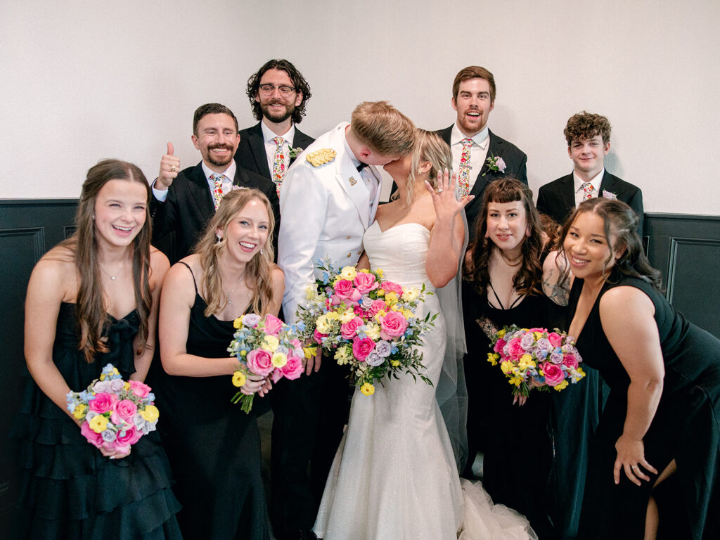 bride and groom kiss and show off ring as wedding party surrounds them