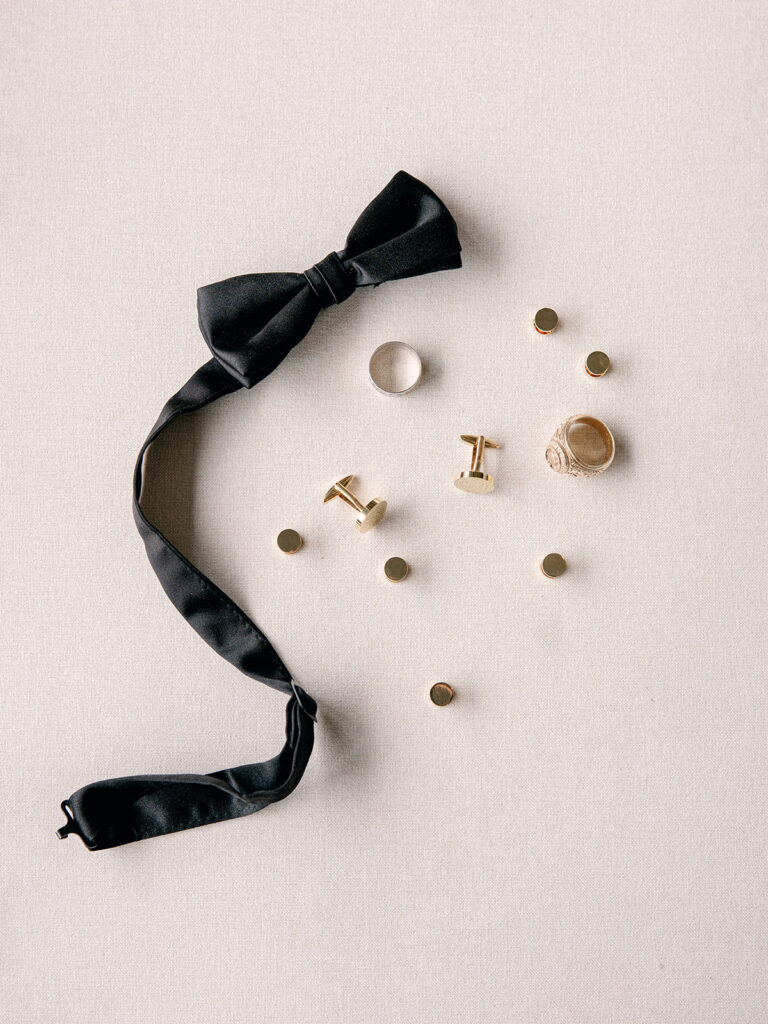 groom's details on a flat lay including his black bow tie, wedding ring and cuff links