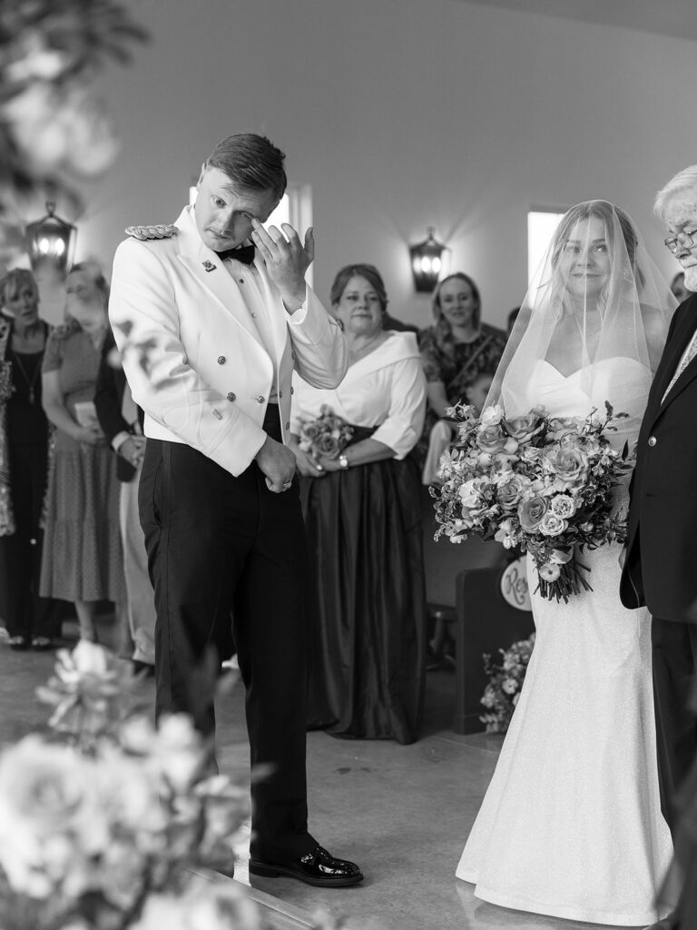 groom wiping away tears as bride stands before him with her dad at the top of the aisle for their wedding at Davis & Grey Farms