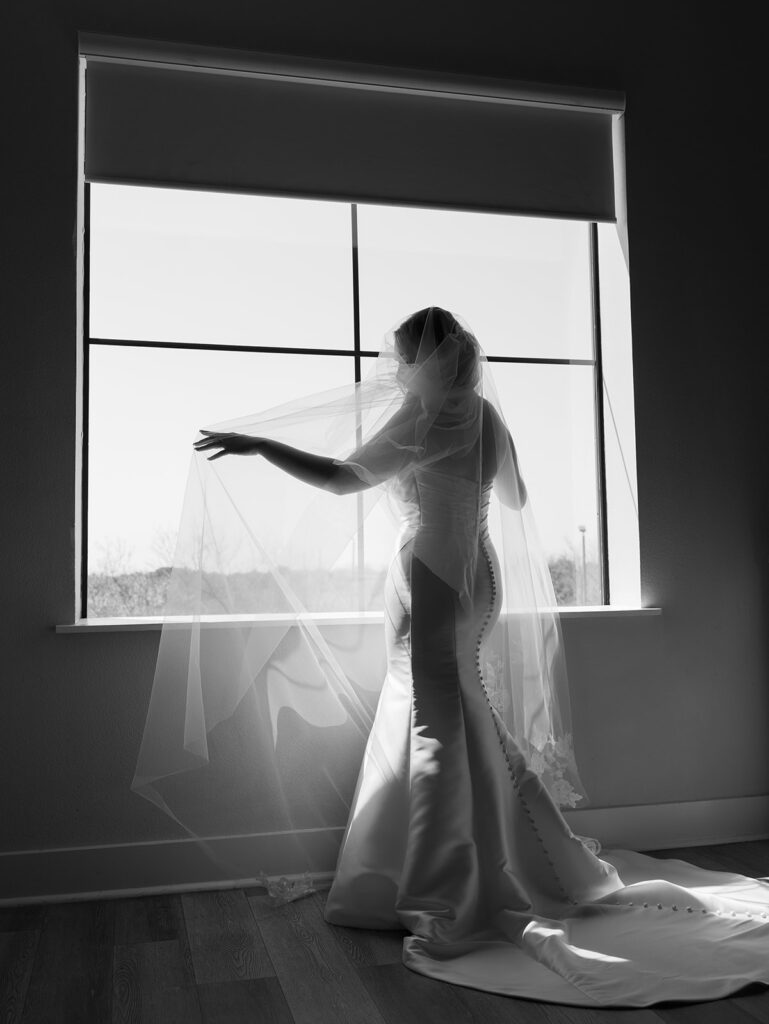 Bride floating her veil in front of a window as she looks like a silhouette 