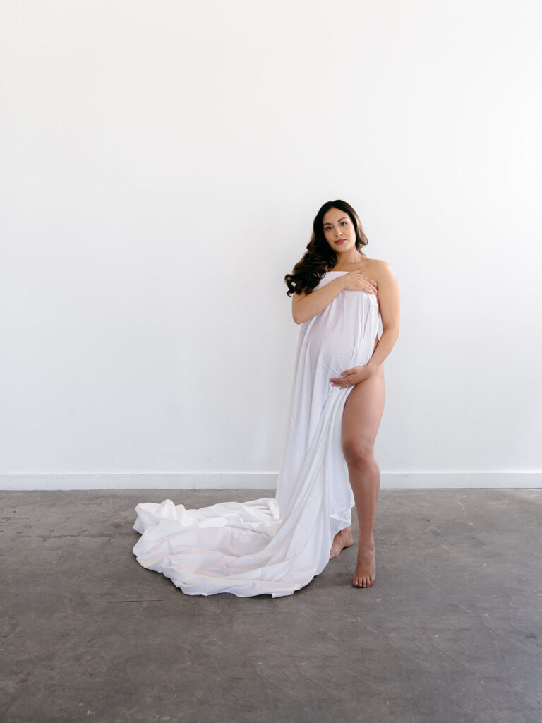 mom holding her pregnant stomach covered in a white bed sheet during a studio maternity session