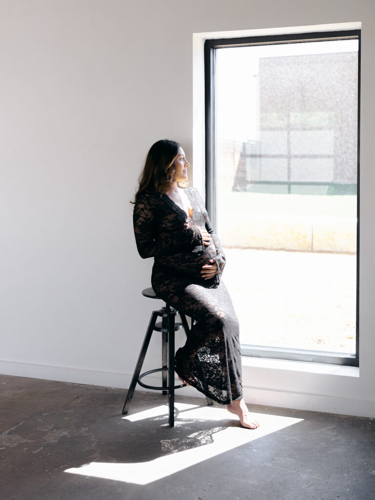 studio maternity session with mom on a stool holding her stomach looking out a window

