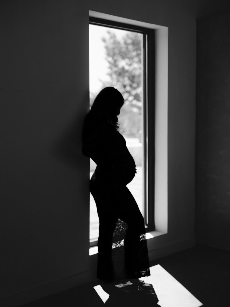 pregnant mom's silhouette standing next to a window looking at her stomach during a studio maternity session