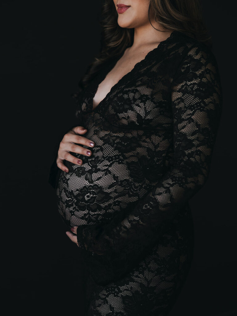 Studio maternity session with a black backdrop and a black lacy dress with mom holding her tummy