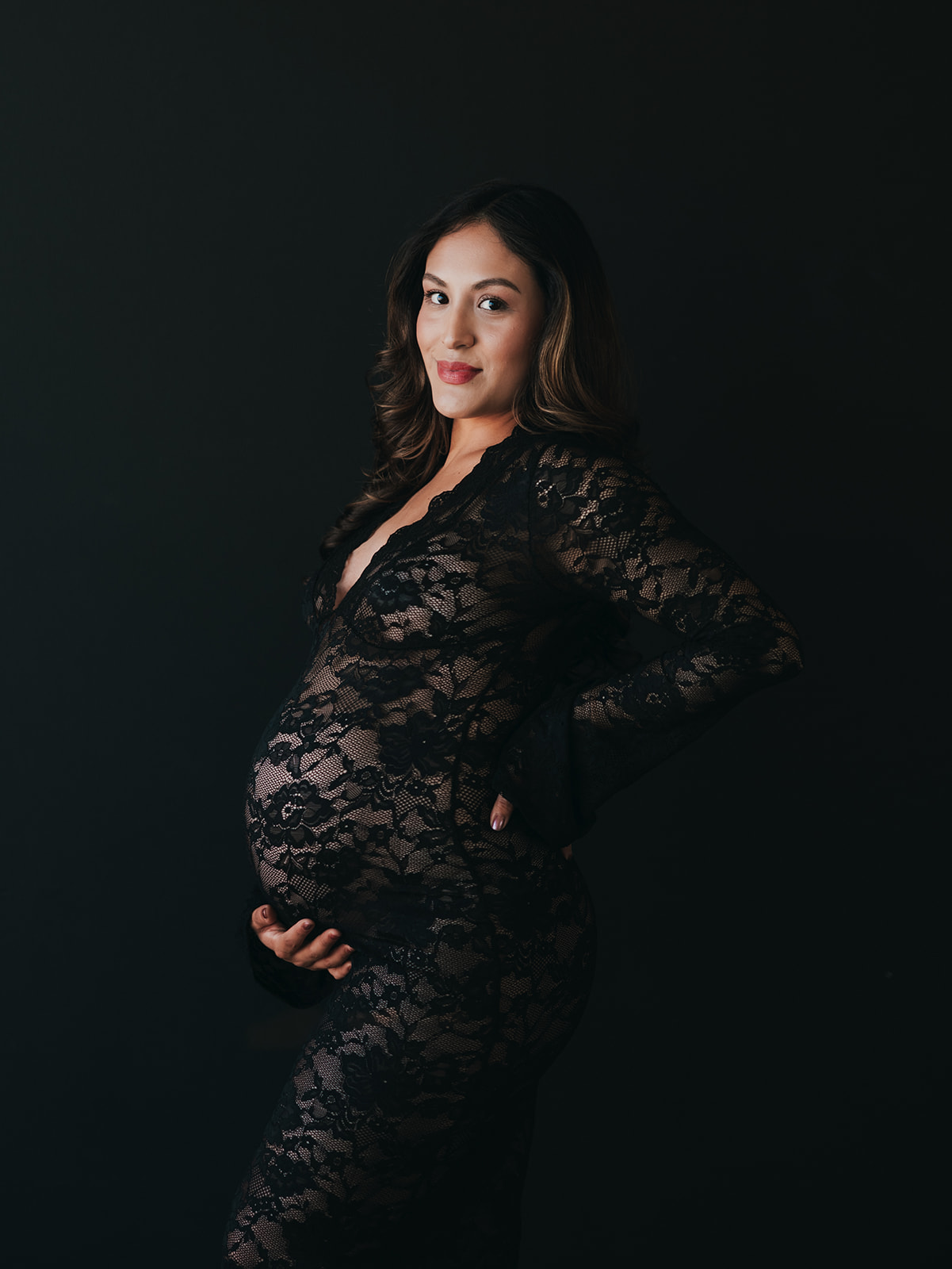 pregnant mom during a maternity studio session wearing a lacy black dress standing in front of an all black background