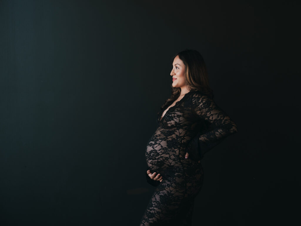 Studio maternity session with a black backdrop and a black lacy dress with mom holding her tummy