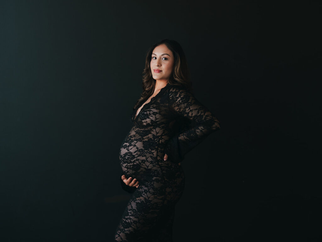 Studio maternity session with a black backdrop and a black lacy dress with mom looking at the camera with a soft gaze
