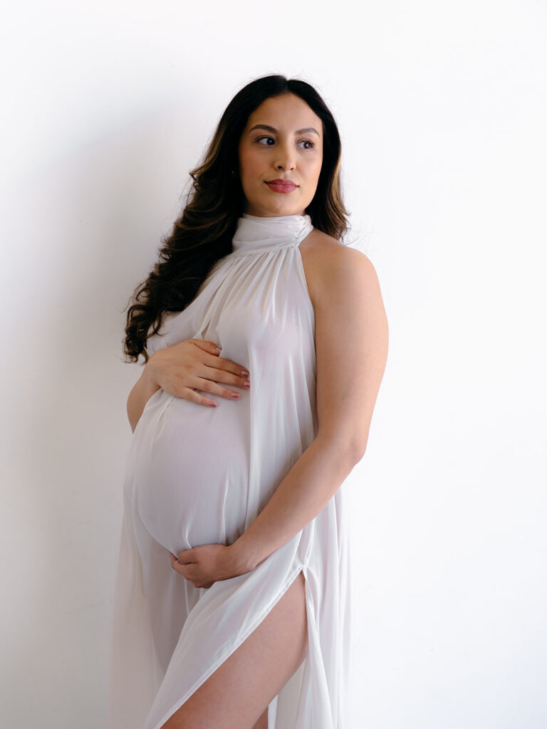 mom holding her pregnant stomach wearing a white flowy gown during a studio maternity session