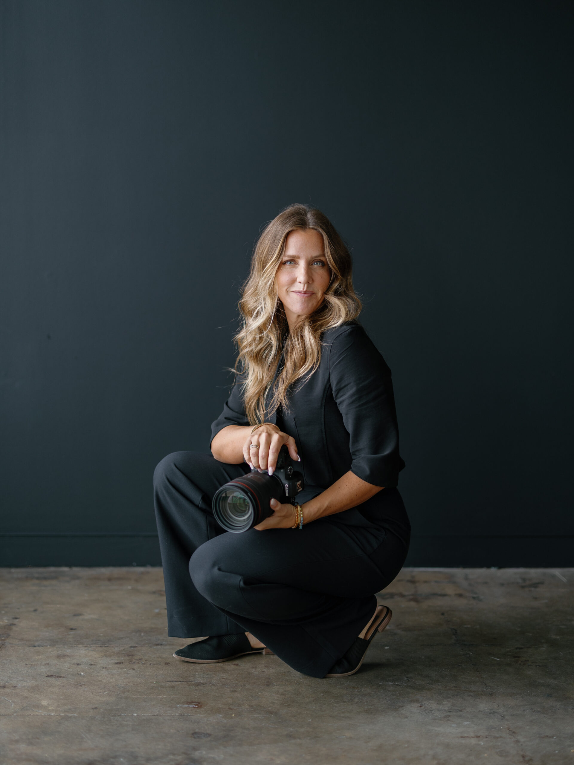 branding photo of Molly Murphy Photography in wedding outfit holding camera on a black wall